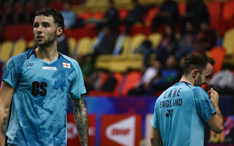 Sean Vendy and Ben Lane are wearing light blue tops with an England flag in the corner. Sean is looking towards the camera and Ben is facing away